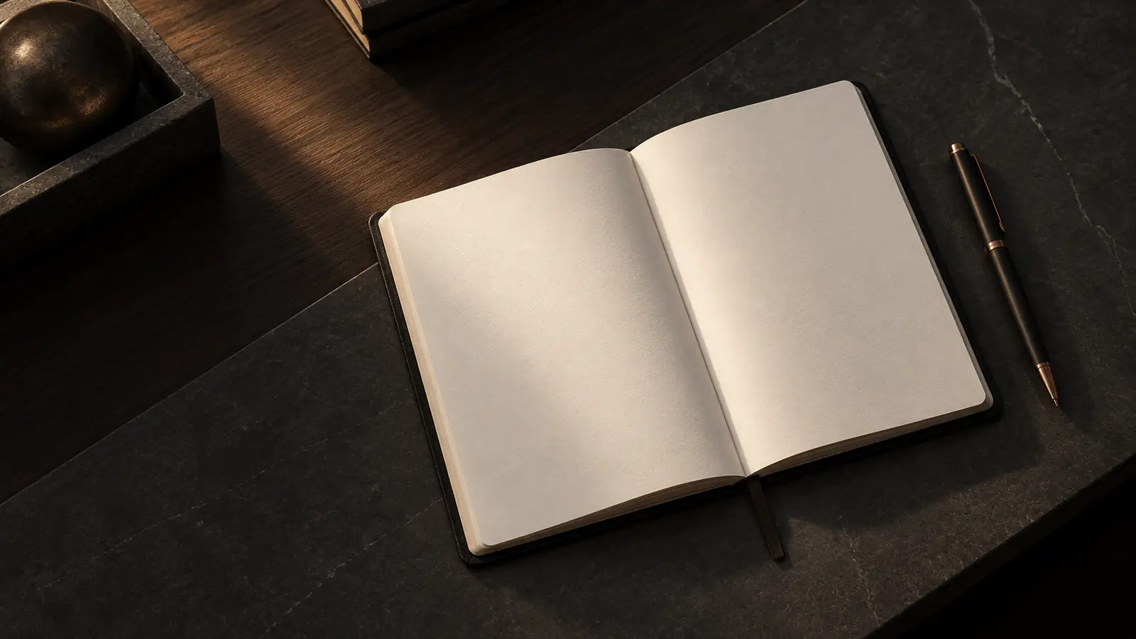 Quaderno aperto con pagine bianche e una penna su un piano in pietra scura.